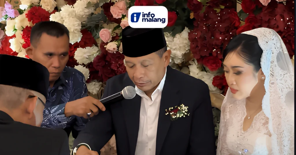 Wali Kota Malang Wahyu Hidayat mengenakan peci hitam, jas hitam, dan kemeja putih saat prosesi ijab kabul, duduk di samping mempelai perempuan berkebaya brokat putih dan berkerudung renda, dengan latar dekorasi bunga merah, putih, dan merah muda.